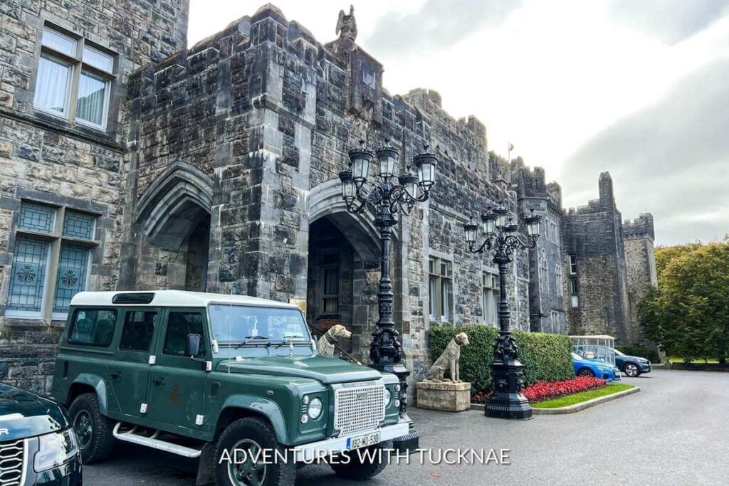 The entrance to Ashford Castle. The Castle is grey stone with square walls and arched entry points. There is an old Range Rover parked by the entrance with ornate lamp posts and stone dog statues on either side of the entryway. 