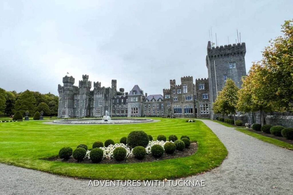 The view from the gardens outside of the Ashford Castle Hotel in Ireland. There is a fountain and shrubs on the lawn in front of the beautiful castle hotel. 
