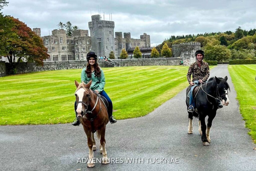 A couple riding horses with Ashford Castle in the background. One horse is brown with a white stripe down his nose, and the other horse is black with a white stripe down his nose. 