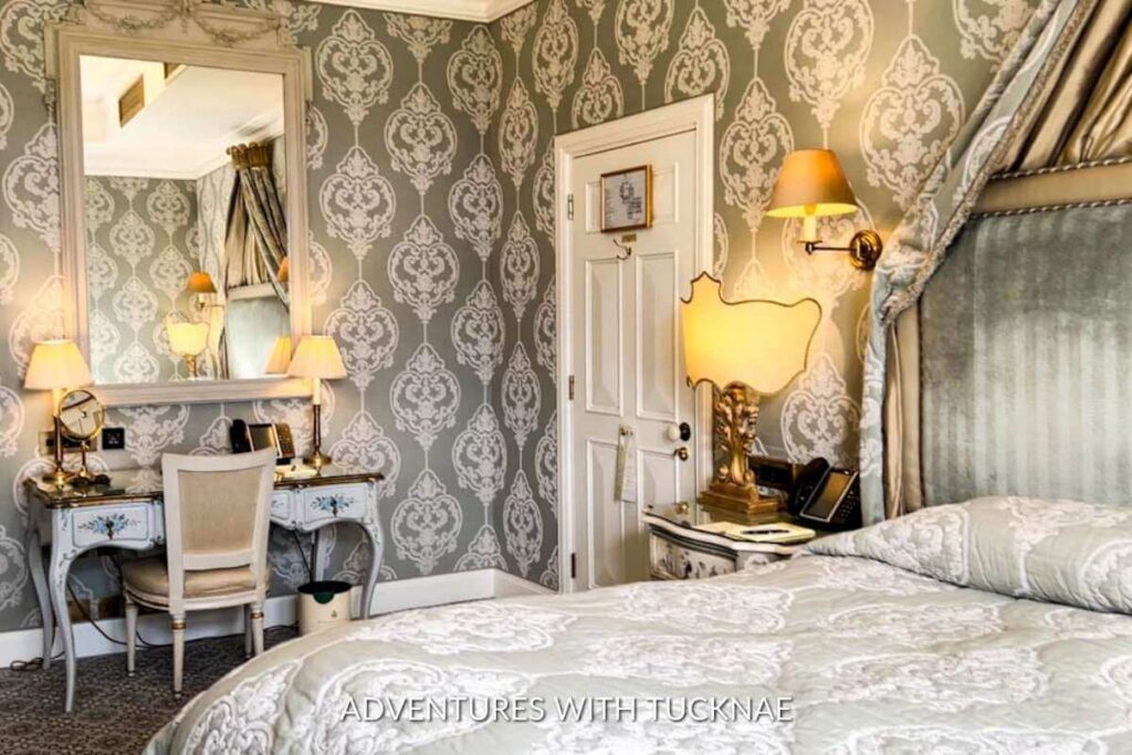 Lake view room in Ashford Castle hotel. The image shows the interior of the room with light green accents. There is beautiful wallpaper covering the walls, a romantic oversized bed, a desk, lamps, and a mirror on the wall.