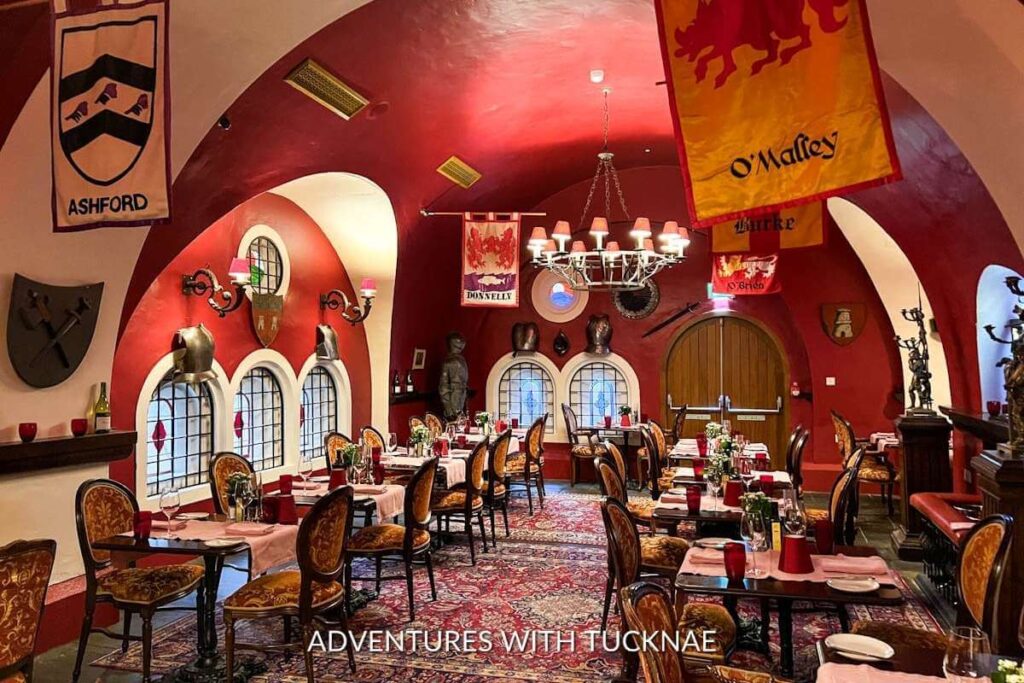 The Dungeon Restaurant in Ashford Castle. The walls are red and there are pendants with family crests hanging from the ceiling. There are tables set throughout the room.