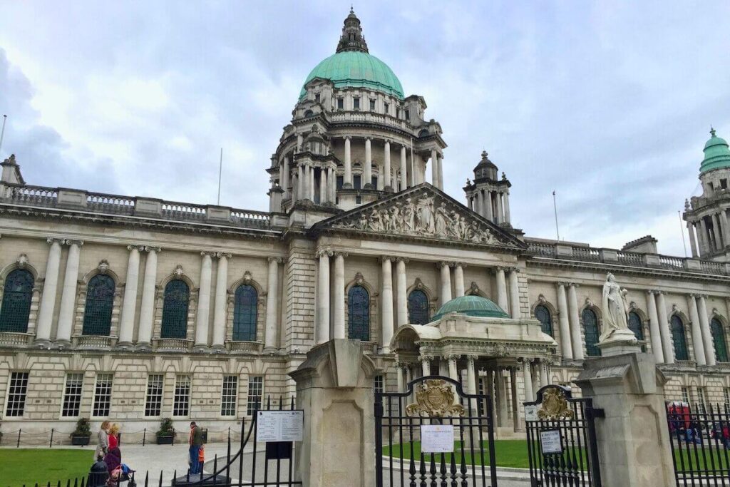 The outside of Belfast City Hall in Northern Ireland. The building is large with pillars, turrets, and decorative elements, and there is a gated entry.