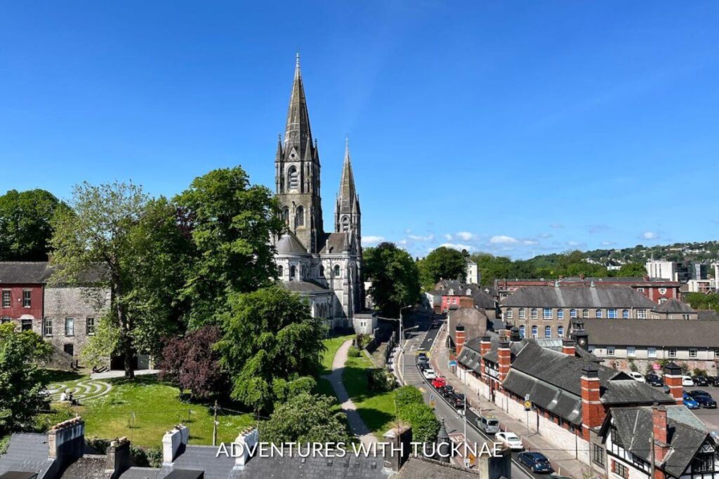 A view of the city of Cork in Ireland. There are houses and small buildings, as well as a tall cathedral with spires. Trees and shrubs surround the area. 
