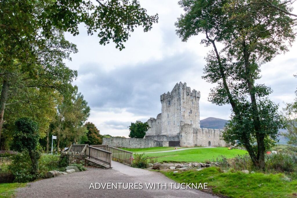 A distant view of Ross Castle in Killarney, Ireland. The castle is a square tower with shorter castle walls surrounding it and green gardens around the base. 