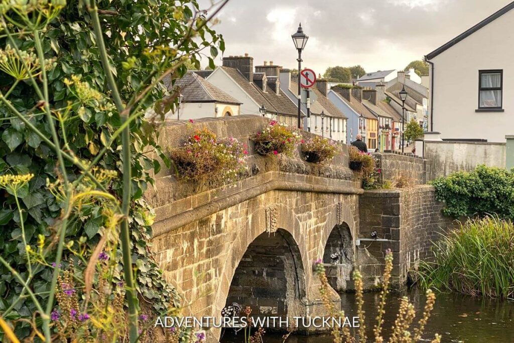 The bridge going to a town in Westport, Ireland. The bridge is stone with two archways underneath and flower boxes on the sides. The town is visible in the distance, with colorful houses lining the road.