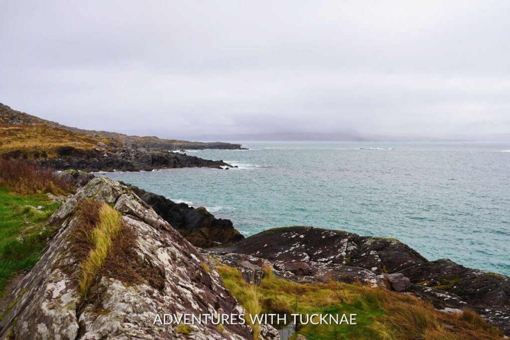 The Wild Atlantic Way coastline in Ireland on a rainy day. There is fog in the distance, with a green and brown shoreline being splashed with bright blue water.
