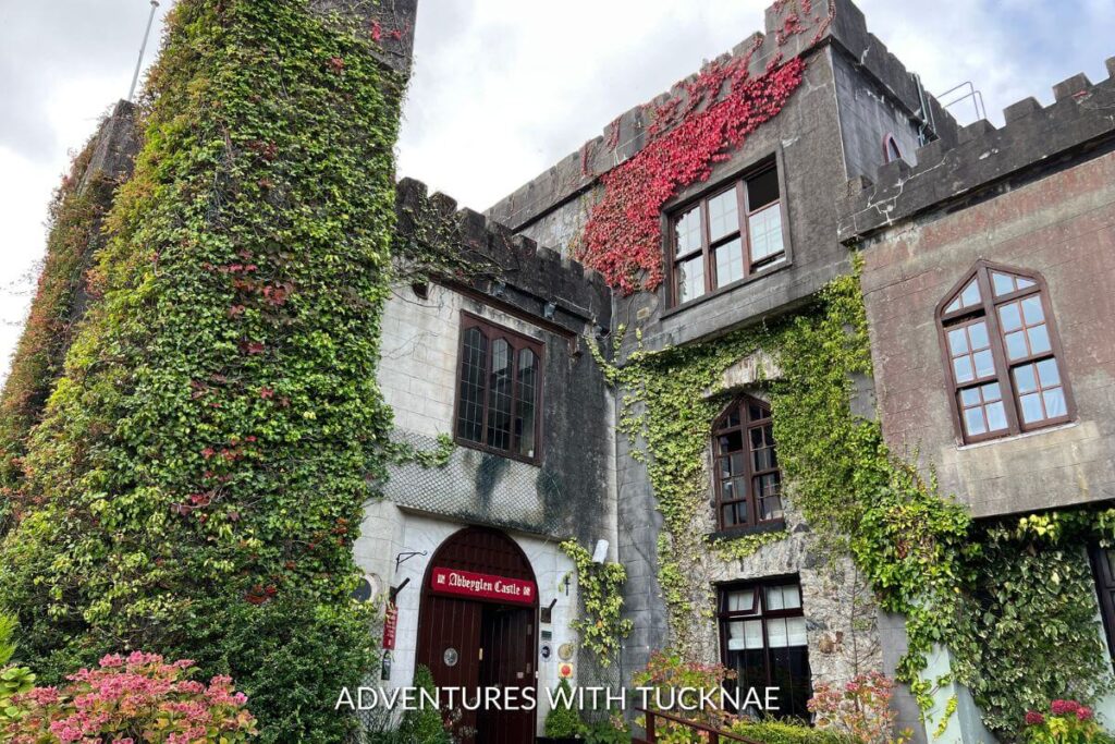 View of the outside of Abbeyglen Castle hotel in Clifden, County Galway, Ireland. There is a red sign reading "Abbeyglen Castle" over the round doorway, and the castle exterior is covered in bright green adn red ivy.