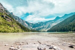 Standing on the edge of Avalanche Lake in Glacier National Park. The sky is bright blue with white clouds, the mountains are green, and the water is murky with large rocks on the edges.