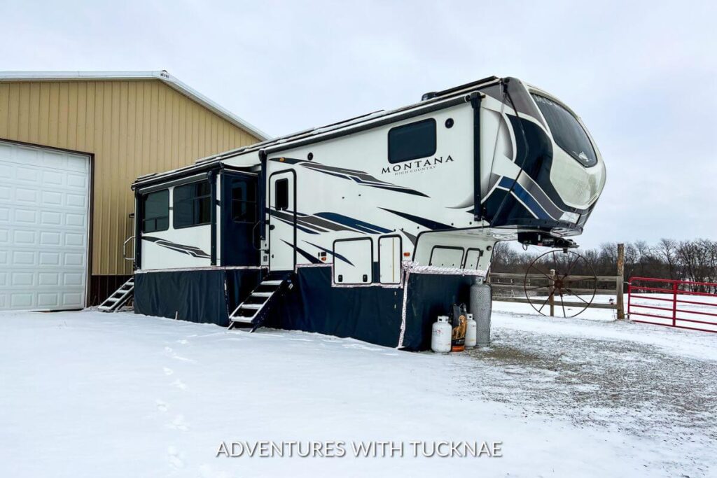 Our 5th wheel RV in winter. The travel trailer has black skirting surrounding it, and the ground is covered in snow.