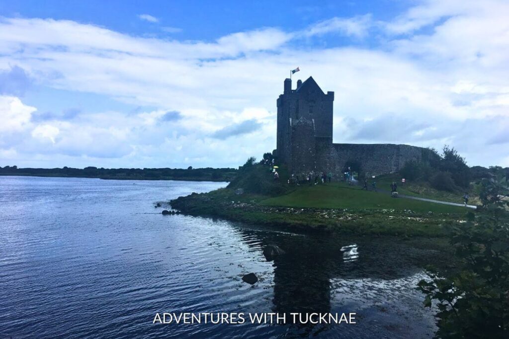 Dungauire Castle reflecting in Galway Bay with the Ireland flag flying above the castle/ The sky is blue and full of whispy white clouds