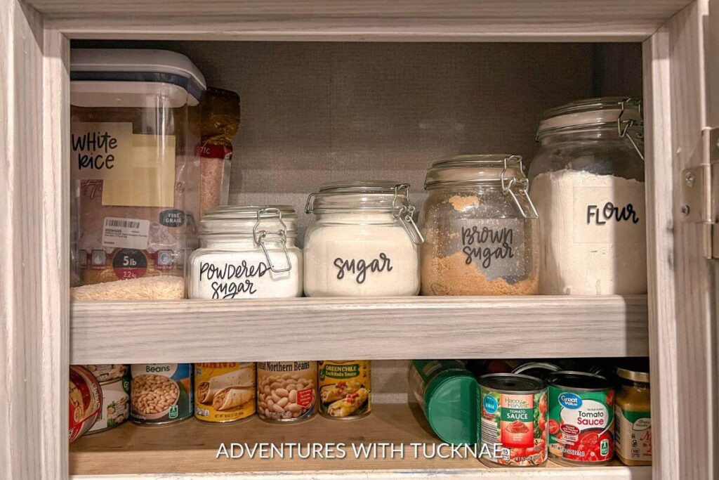 Glass jars in our RV pantry with labels reading white rice, powdered sugar, sugar, brown sugar, and flour. There are also cans of food on the next shelf down. 