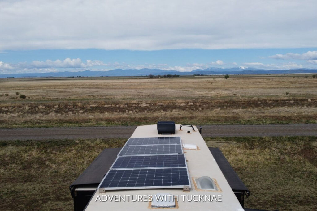 The view from above of an RV roof shows the solar panels on top of the RV. There is a large field in front of the RV with mountains in the distance.