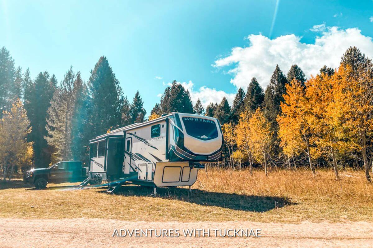 A Keystone Montana High Country 335BH fifth-wheel RV boondocking in the fall. It is surrounded by orange and yellow trees and a black pickup truck is in the background.