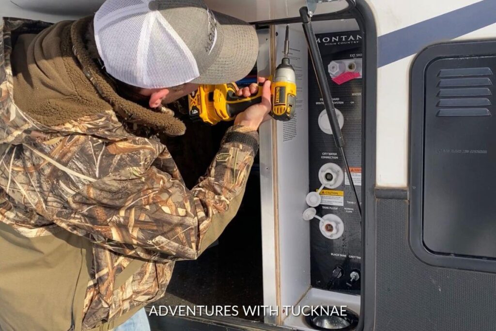 Tucker removing the original water panel in our RV to add our new RV water panel. He is wearing a white and grey ball cap and a camo coat
