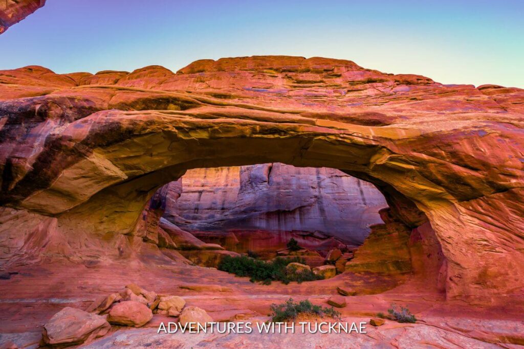 The sun setting over Tower Arch in Arches National Park. The sunset is casting purple and pink hues over the orange arch.