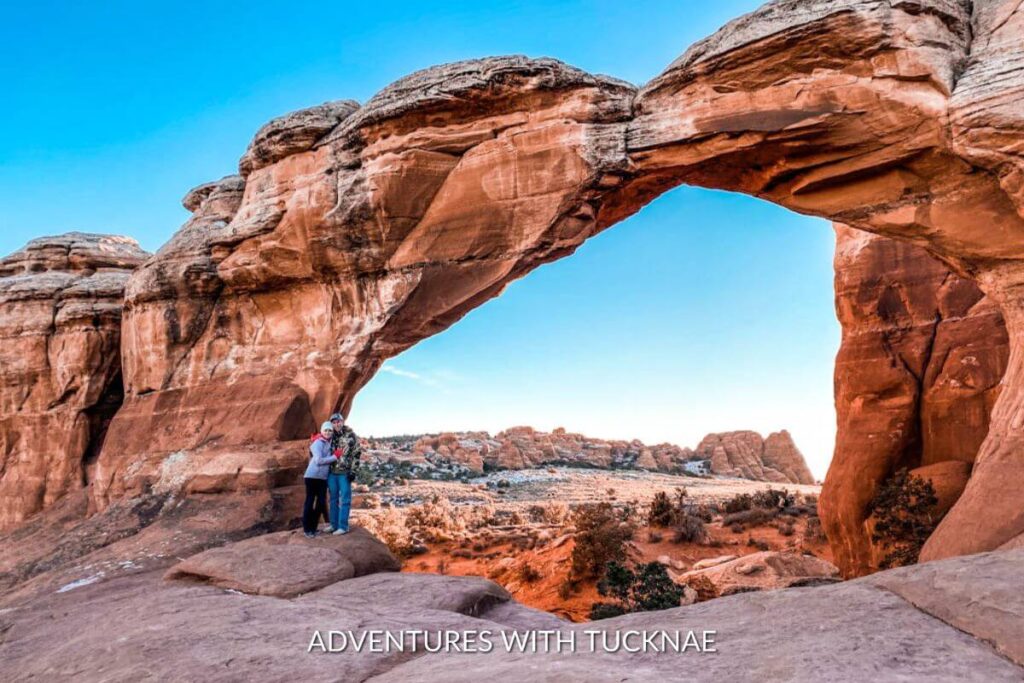 A couple standing underneath of Broken Arch during a hike in Arches National Park.