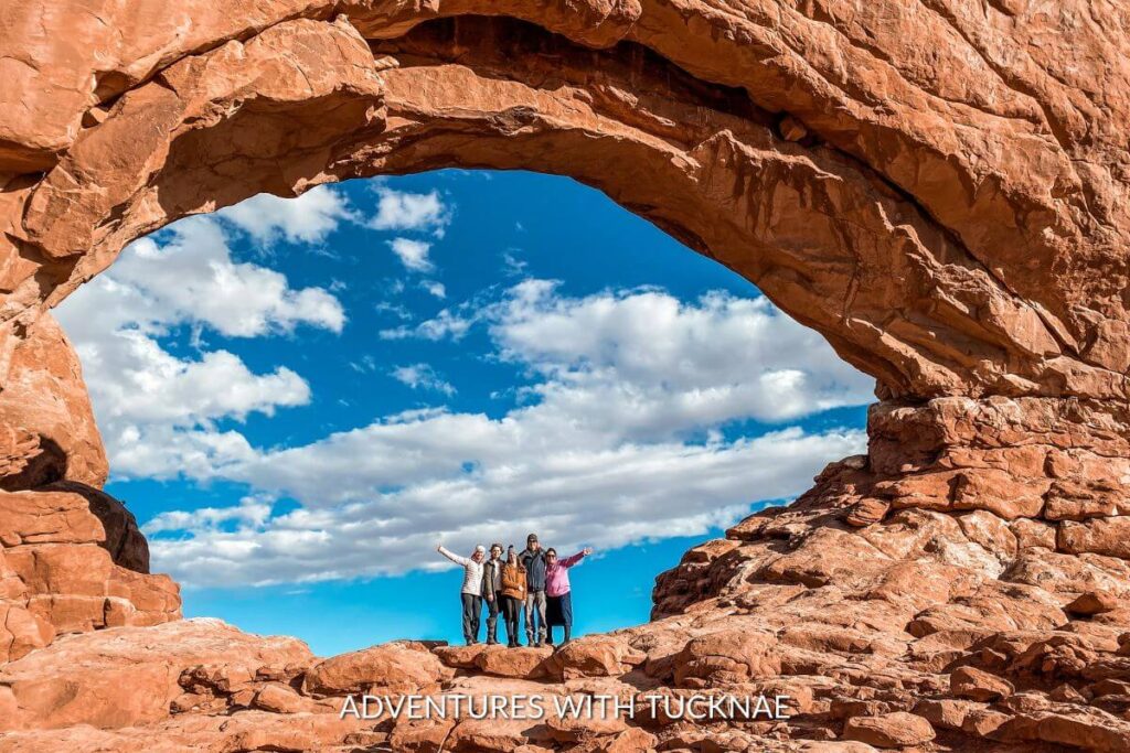 A group of people standing inside of the North Window Arch in Arches National Park. The arch is orange sandstone and the sky behind is bright blue with white clouds. 