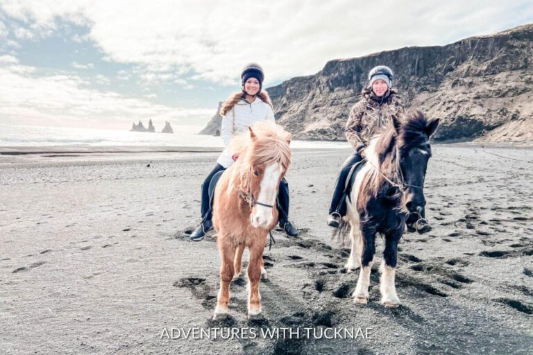 A man and a woman horseback riding on a black sand beach near Vík, Iceland.
