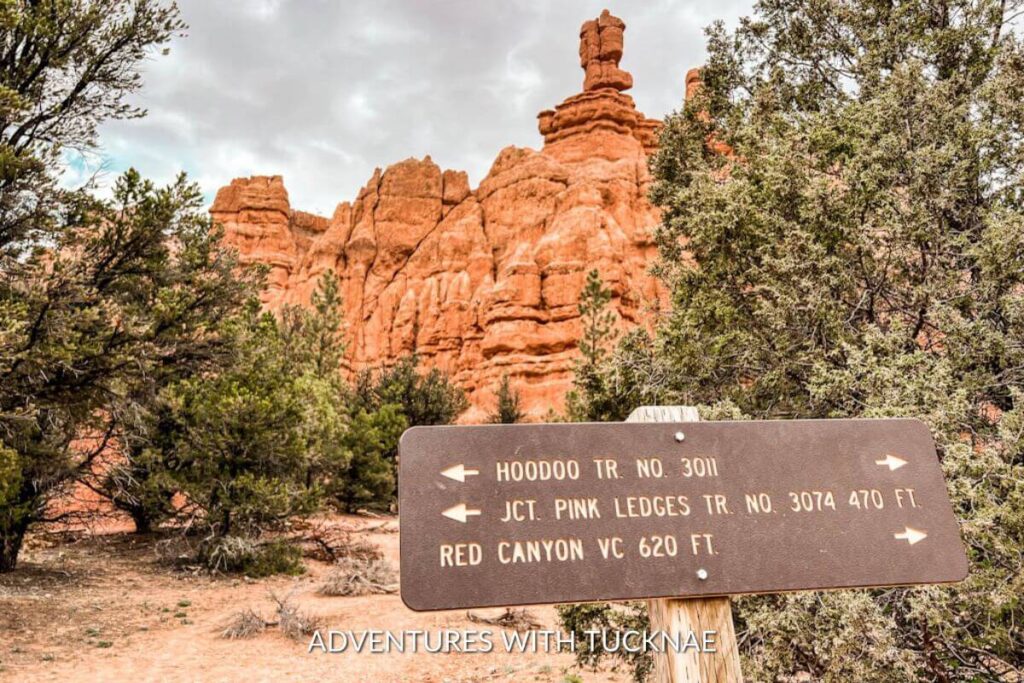 A trail sign showing the hikes available in Red Canyon in Dixie National Forest in southern Utah. The sign is brown and shows the information for 3 hikes. Red hoodoo formations are visible in the background.