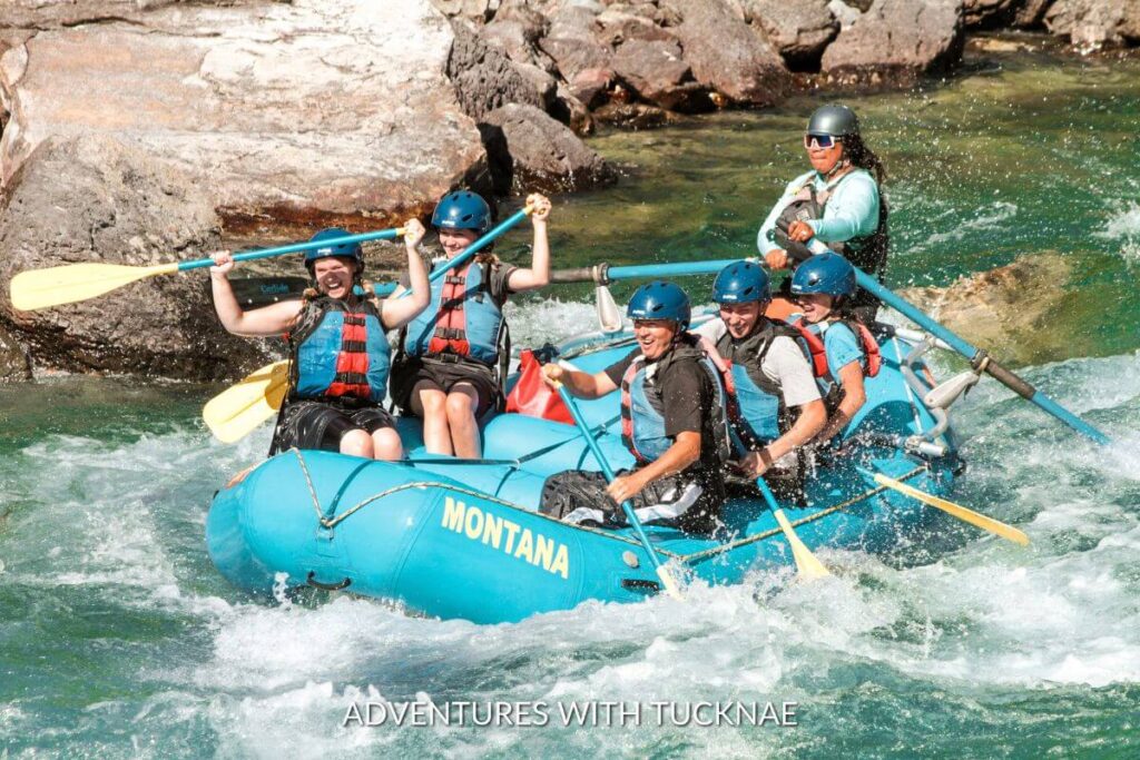 A group of people white water rafting in a blue raft with yellow ores. The people are laughing and having a great adventure.