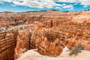 The view of the hoodoos in Bryce Canyon National Park from above. The hoodoos and rock walls are orange and the sky above is bright blue with white clouds.