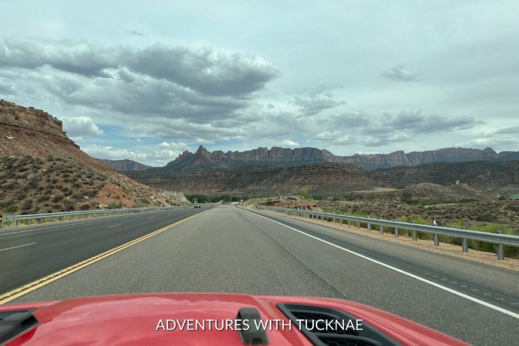 Image of a scenic drive in southern Utah. The hood of a red vehicle is visible and the picture is taken through the front windshield looking towards the road. There are mountains visible in the distance