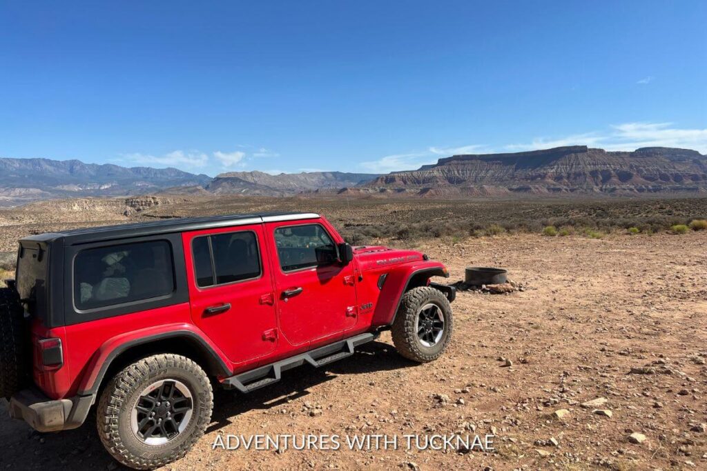 Jeep camping outside of St George, Utah. The Jeep is red and black and there are mountains surrounding the area in the distance.