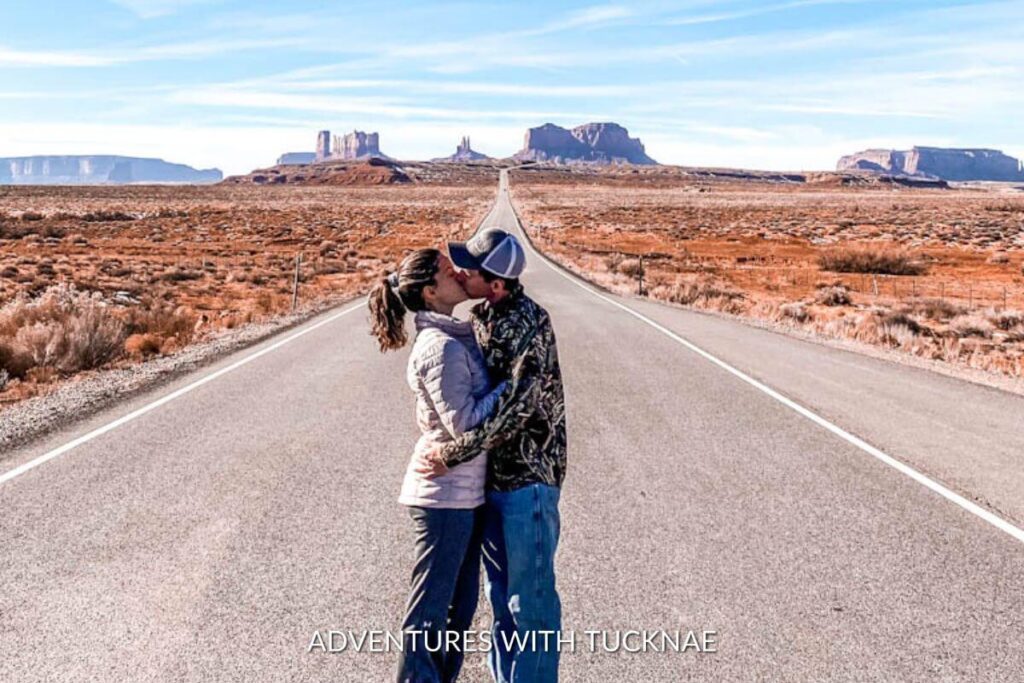 A couple standing in the road kissing with Monument Valley sprawling out in the distance behind them.