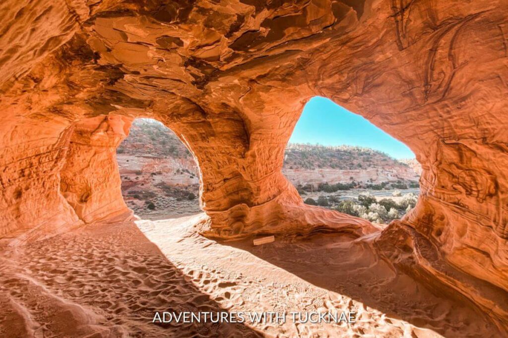 An inside view of the Moqui Caves near Kanab, Utah. There are two openings on the edge of the caves. The walls and ceiling are orange sandstone and the floor is sandy.