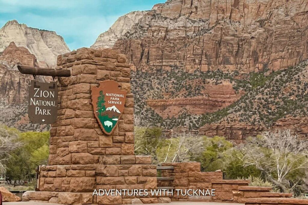 The entrance sign for Zion National Park with a beautiful view of the park in the background. 