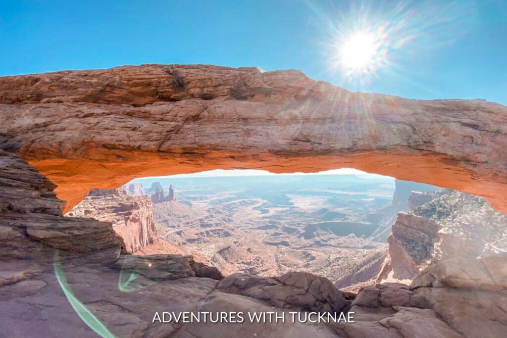 A natural archway in Canyonlands National Park with the canyon shown down below and the sunshine peeking behind the arch. 