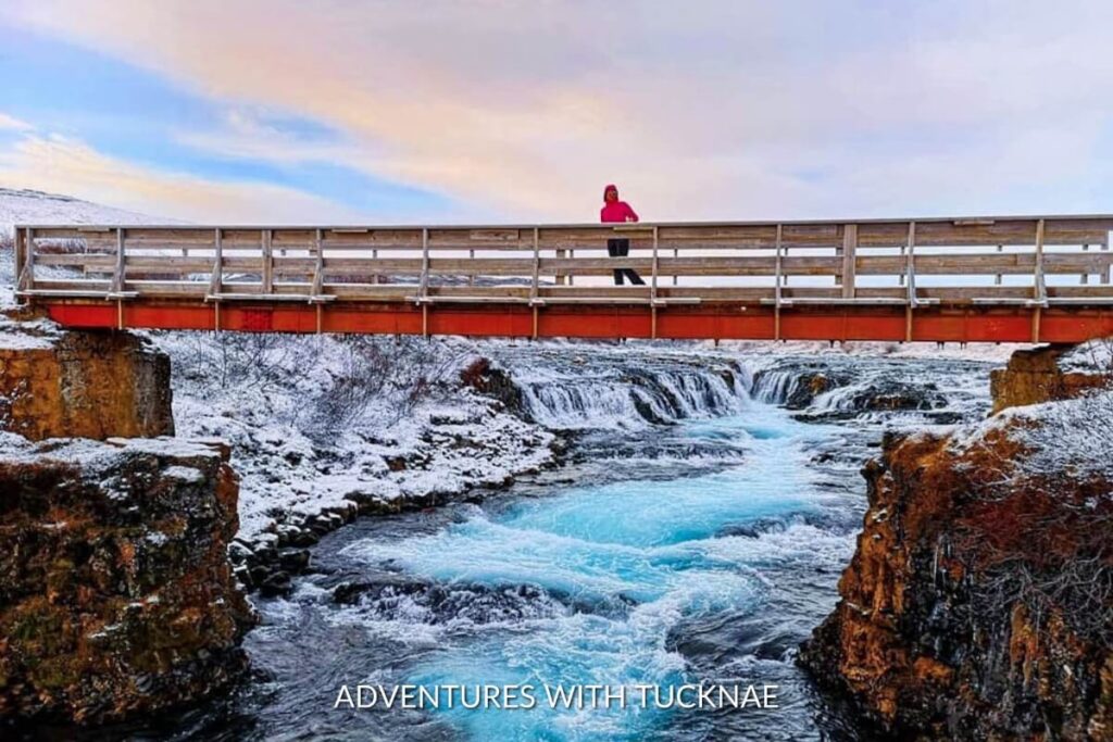 A snowy scene of Brúarfoss waterfall in Iceland with a man standing above it on a bridge