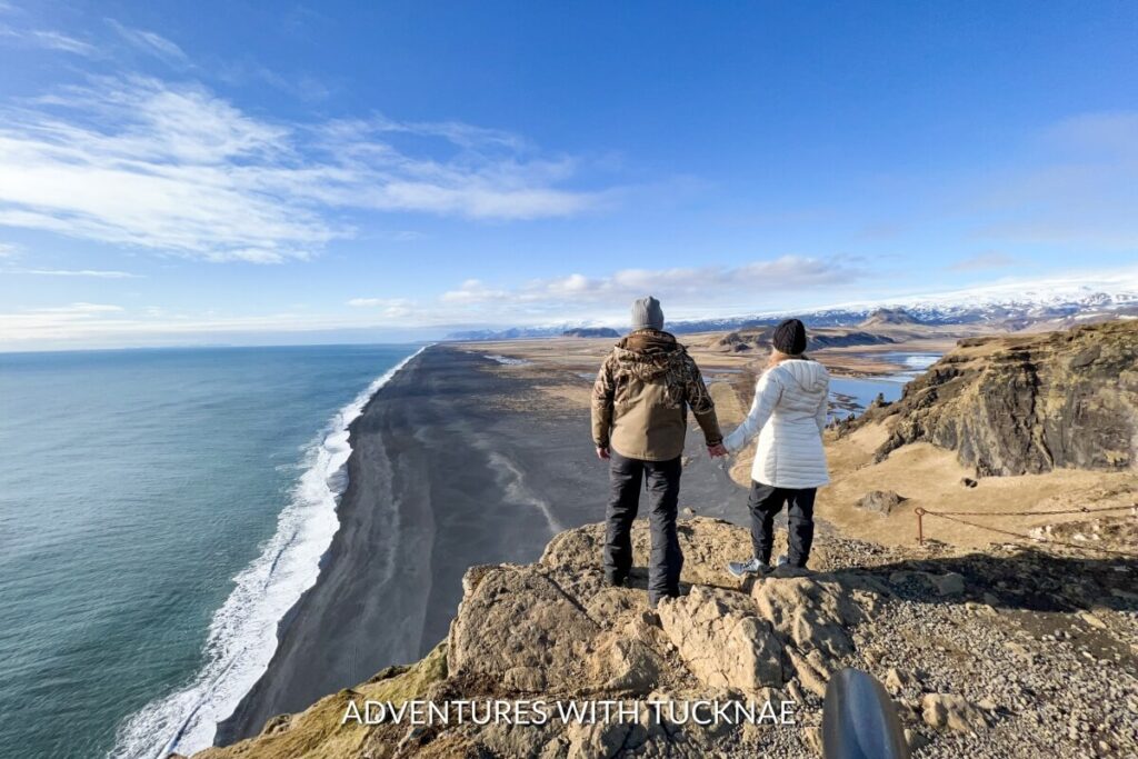A couple standing on the edge of a cliff in Iceland looking out over the black sand beach below