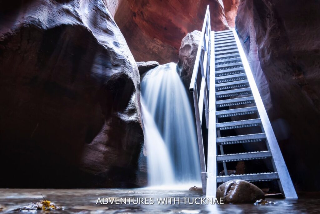Water cascading down a small fall with a metal ladder alongside, within the shadowy confines of Kanarra Creek Canyon, Utah.
