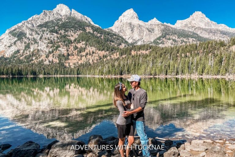 A couple shares a romantic moment on the shores of Taggart Lake with the Teton Range reflected in the water, on the Taggart Lake and Bradley Lake Loop hike in Grand Teton National Park.