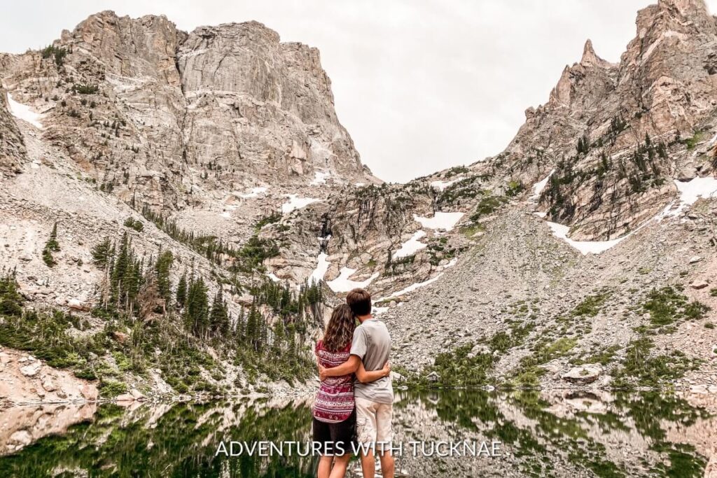 A couple embracing while gazing at the tranquil Emerald Lake surrounded by rugged peaks, a picturesque spot on the bucket list national park hike in Rocky Mountain National Park.