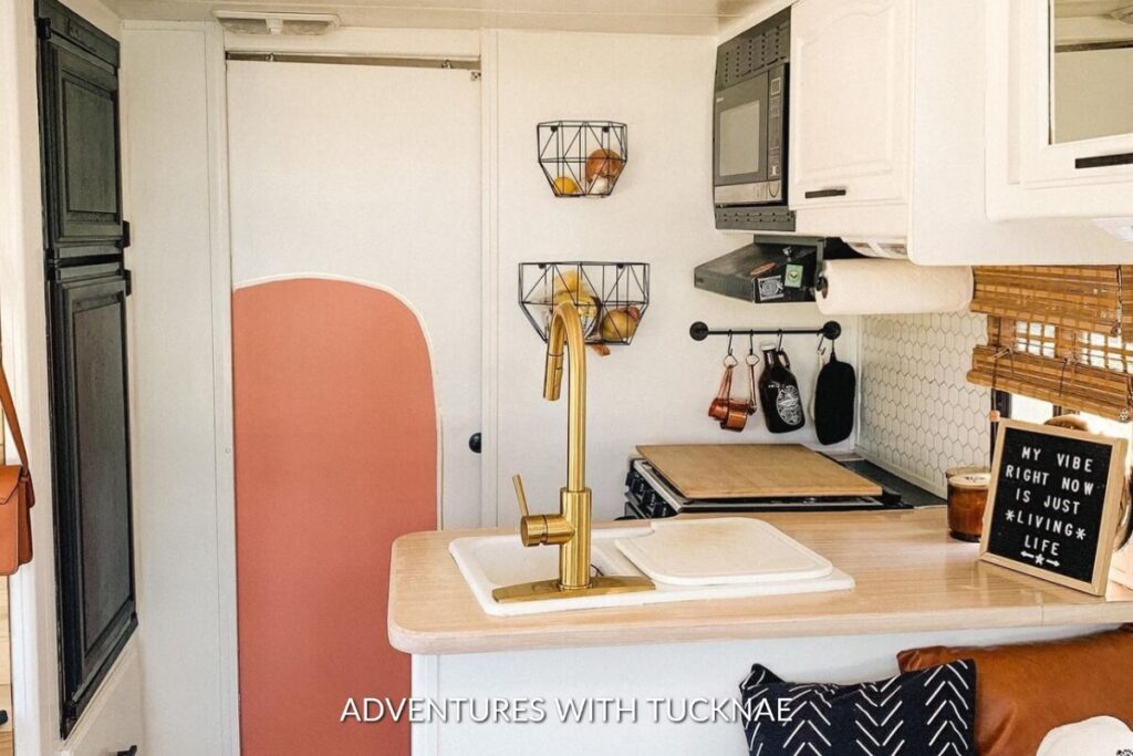 A stylish RV kitchenette with a golden faucet, white countertops, a microwave, and a sign reading 'My vibe right now is just living life', adding a personal touch.