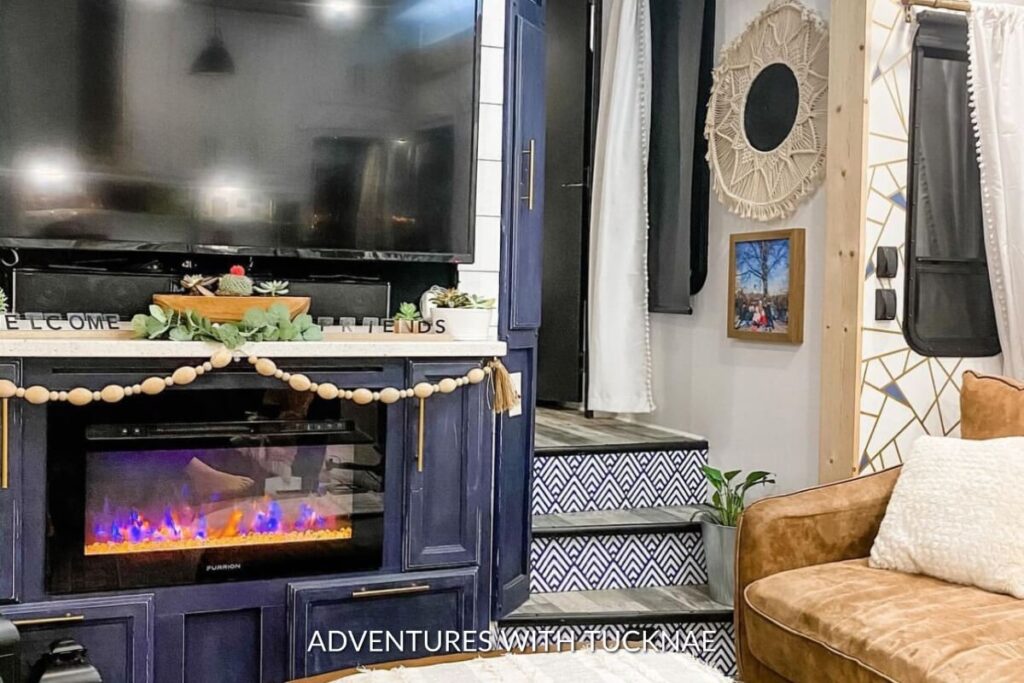 A stylish RV living area with a black entertainment center and electric fireplace below a 'Welcome Friends' sign, adjacent to a cozy sofa corner with a macrame wall hanging.