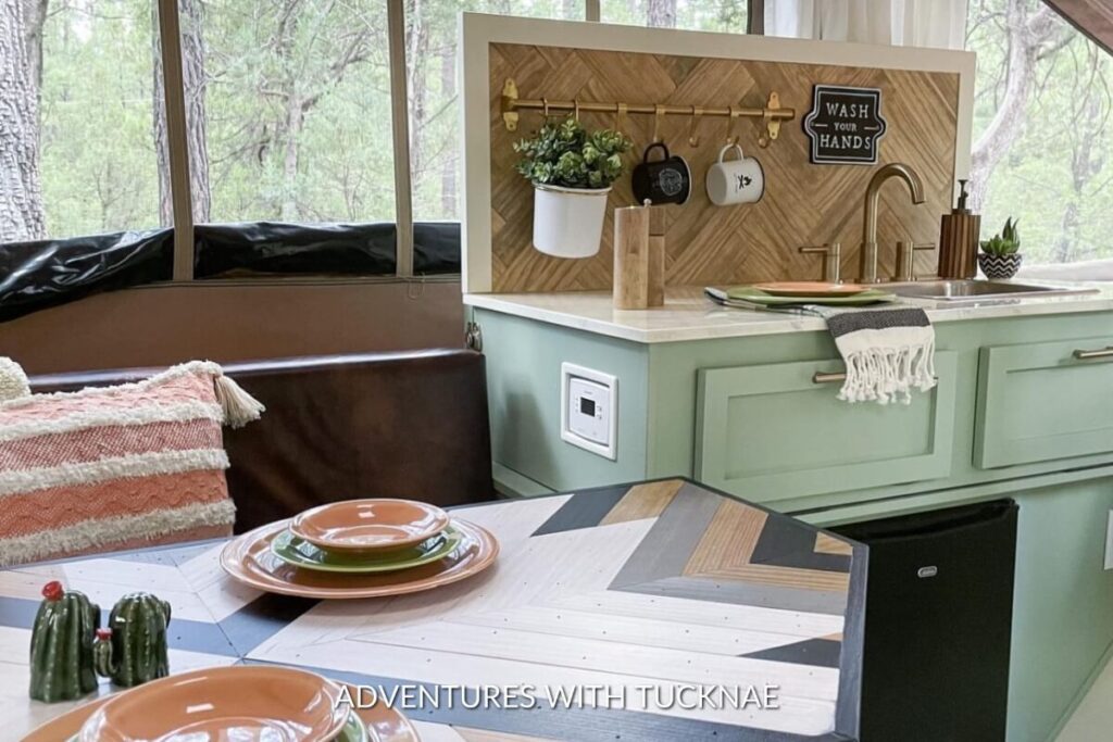 A fresh and natural RV dining nook with mint green cabinetry and a geometric backsplash, featuring a wood dining table set with orange plates and a window view of the forest.
