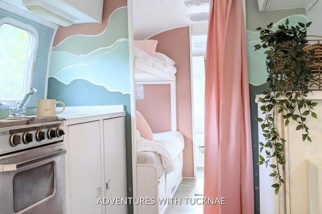 A serene RV sleeping area with ocean wave wall art above a window, a pink bunk bed alcove, and a calming color palette, complete with hanging plants and natural light.