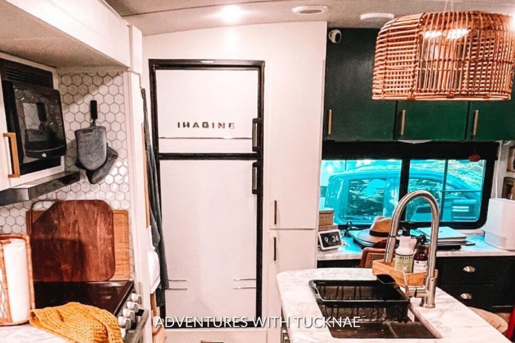 A chic RV kitchen corner with hexagonal tile backsplash, a white fridge, and green upper cabinets, illuminated by a unique wicker lampshade.