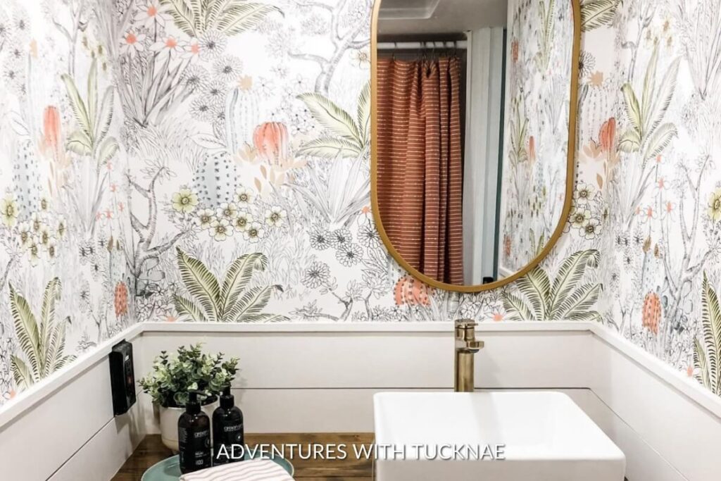 A botanical-themed RV bathroom with a vibrant floral wallpaper, a circular mirror, a modern white sink, and natural wood accents creating a refreshing space.