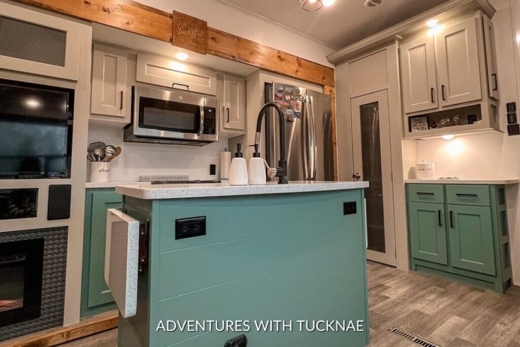 A spacious RV kitchen featuring teal lower cabinets, a large stainless steel fridge, and a central island offering a homey feel.