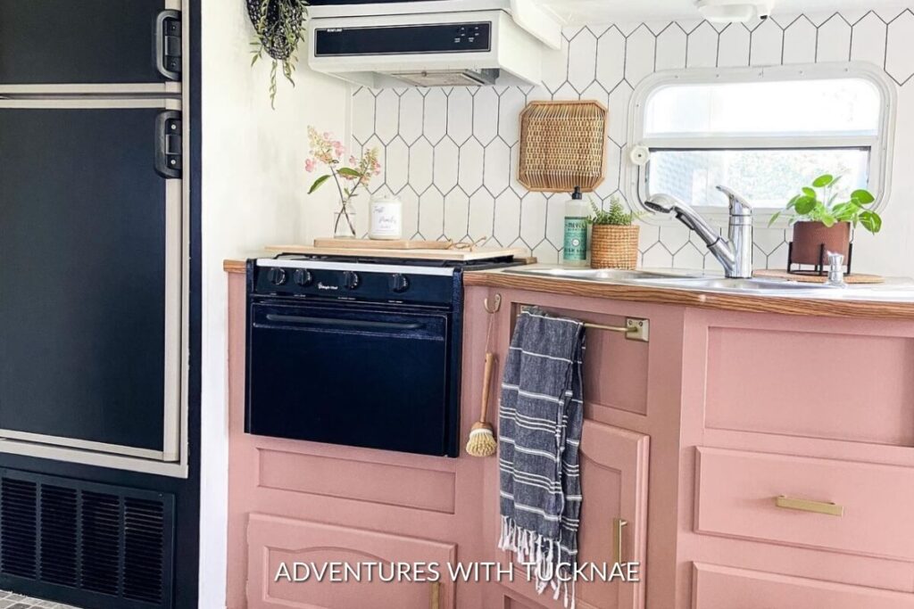 A chic RV kitchen featuring a black stove and oven, with pink cabinets and a white hexagonal tile backsplash, complemented by wicker lampshade and a sink with a potted plant.