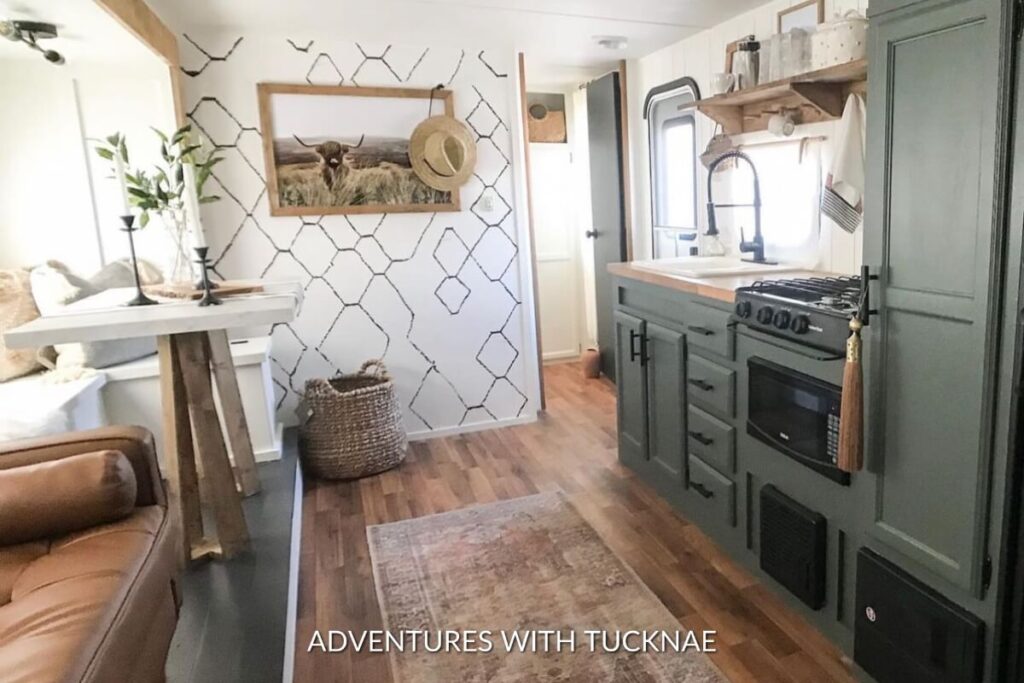 Rustic and cozy RV interior with a warm leather sofa, a white drop-leaf table under a window with a hexagonal pattern accent wall, and a kitchen area featuring green cabinetry and modern appliances.