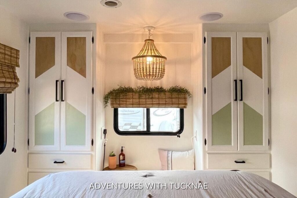 Elegant RV bedroom with a chandelier, white custom cabinets featuring geometric designs, a window with a hanging greenery planter, and a cozy bed with white linens.