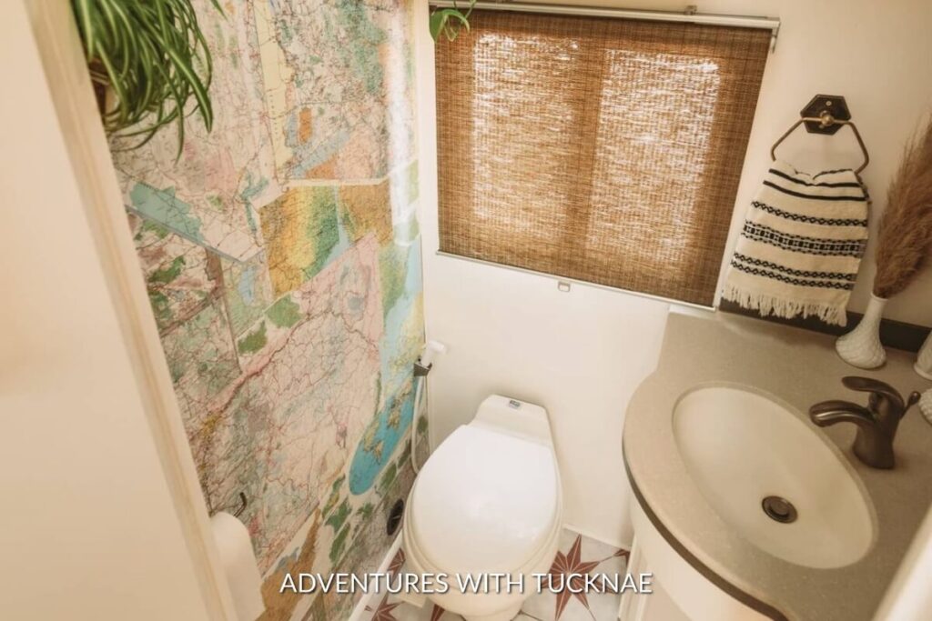 Compact RV bathroom with a map-themed shower curtain, bamboo window shade, white sink, and toilet, enhanced by a decorative striped towel and a potted plant.