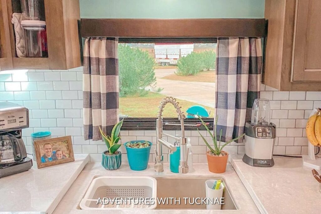 A sunny RV kitchen window dressed with checkered curtains, featuring a white sink, turquoise kitchen accessories, and a photo frame on the countertop.