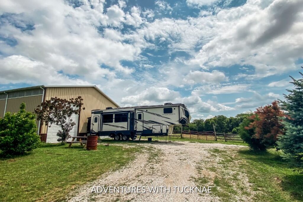A spacious fifth-wheel trailer parked beside a metal storage building, under a sky filled with fluffy clouds, indicating a serene moochdocking setup.