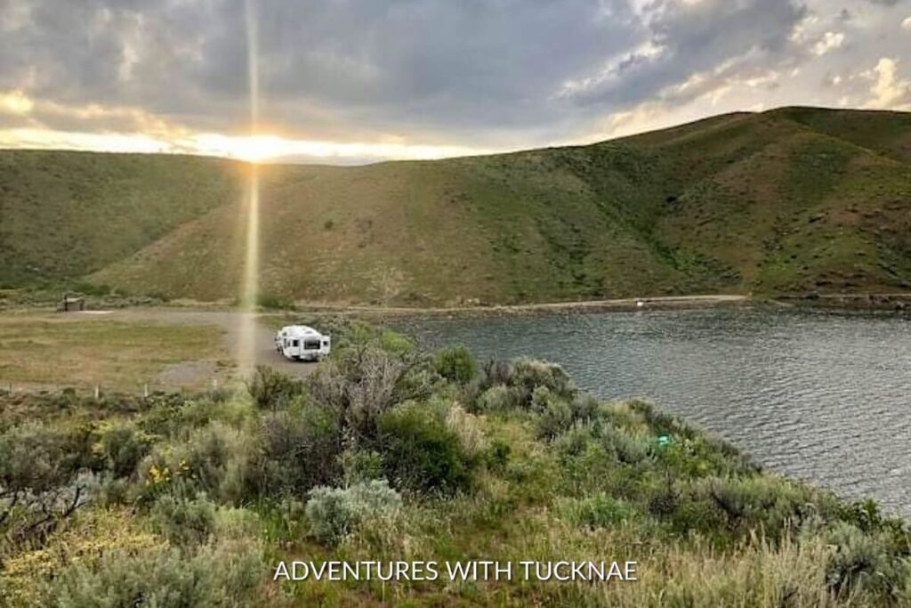 Travel trailer parked on a gravel lot by a serene lake at sunset, with sunbeams piercing through hills in the background, perfect for a boondocking adventure.